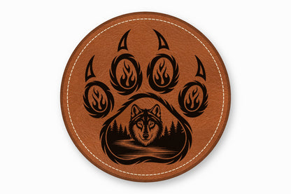 Round brown leather coaster with white stitched edge, featuring a black engraved bear paw and wolf forest scene.