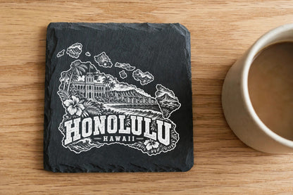 Square black slate coaster engraved with a white Honolulu Hawaii skyline design next to a coffee mug.