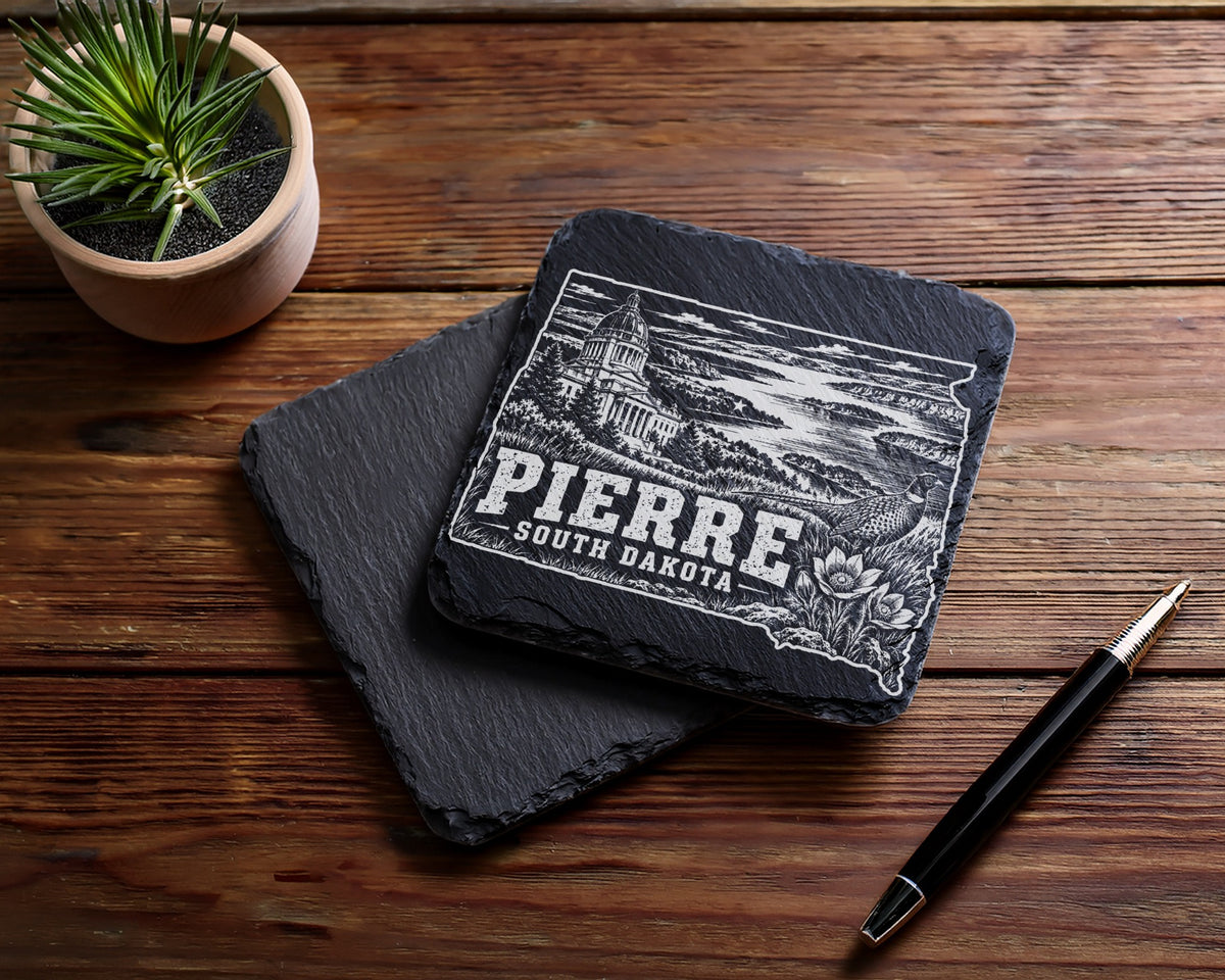 Two square black slate coasters on a wooden table, with an engraved design featuring Pierre, South Dakota.