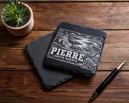 Two square black slate coasters on a wooden table, with an engraved design featuring Pierre, South Dakota.