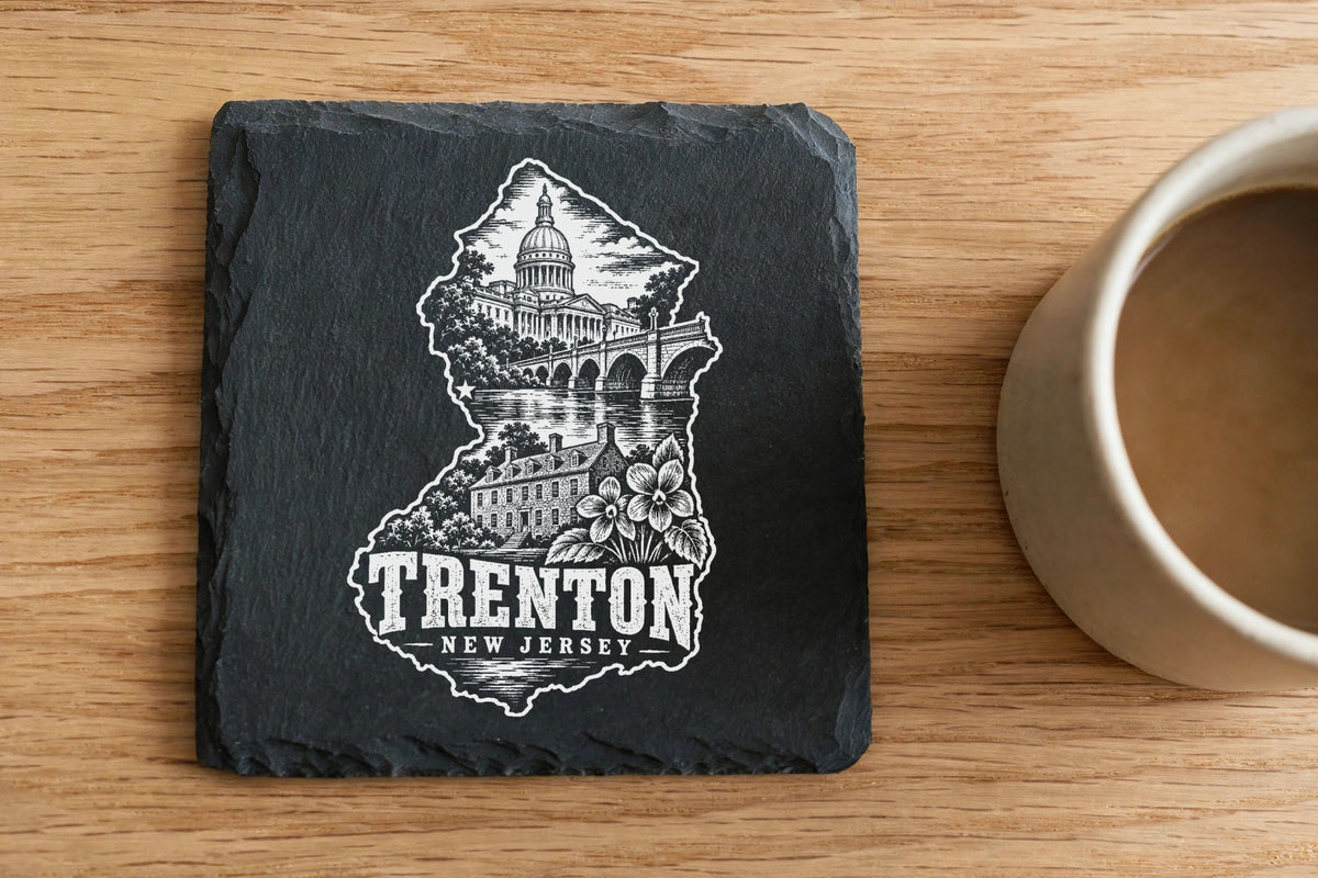 Square black slate coaster engraved with a Trenton, New Jersey skyline and text next to a coffee mug on wood.