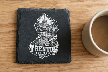 Square black slate coaster engraved with a Trenton, New Jersey skyline and text next to a coffee mug on wood.