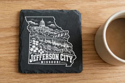 A square black slate coaster engraved with a map outline, landmarks, and the text Jefferson City Missouri rests on a wooden table beside a coffee mug.