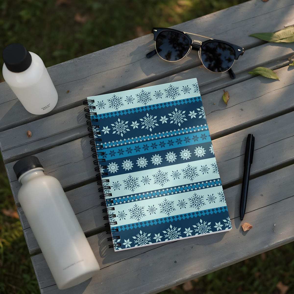 Spiral notebook with blue snowflake pattern, sunglasses, pens, and water bottles on a wooden surface.
