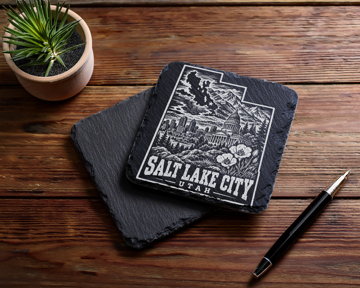 Two square black slate coasters on a wooden table, with Salt Lake City engraved on the top one.