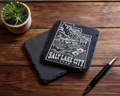 Two square black slate coasters on a wooden table, with Salt Lake City engraved on the top one.
