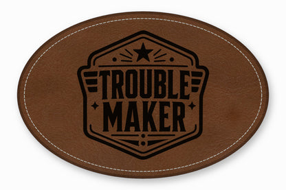 An oval brown leather patch featuring white stitching along the edge and a black printed emblem with the text Trouble Maker.