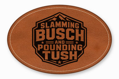 Oval brown leather patch with white stitched border, featuring a black emblem with text reading Slamming Busch and Pounding Tush.