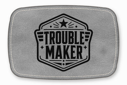 A rectangular grey leather patch with white stitching featuring a black Troublemaker emblem.