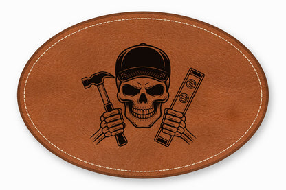 An oval brown leather patch with white stitching featuring a black skull wearing a cap and holding a hammer and level.