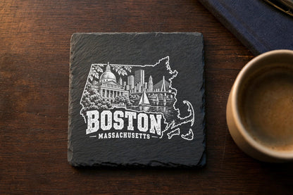 A square slate coaster engraved with a Boston, Massachusetts cityscape inside the state outline on a wooden table.
