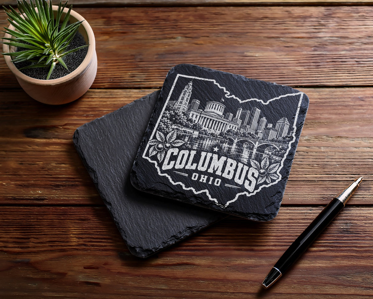 Two square black slate coasters on a wooden table, one engraved with Columbus, Ohio skyline inside the state outline.