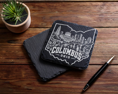 Two square black slate coasters on a wooden table, one engraved with Columbus, Ohio skyline inside the state outline.