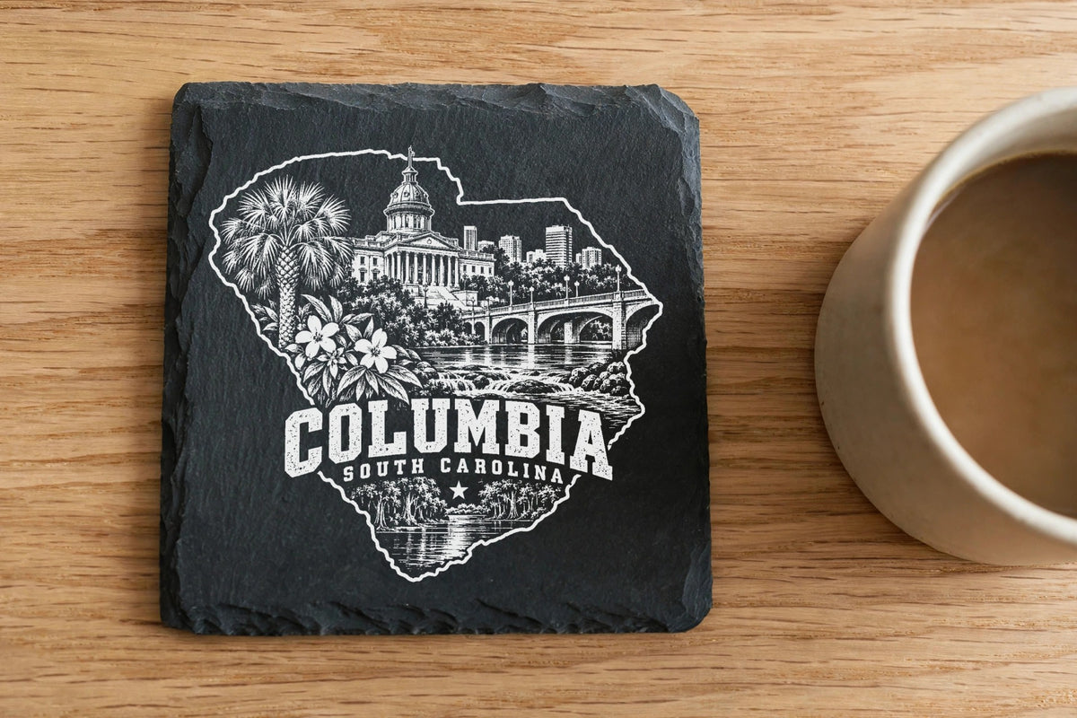 A square slate coaster engraved with a Columbia, South Carolina skyline rests on a wooden table beside a coffee mug.