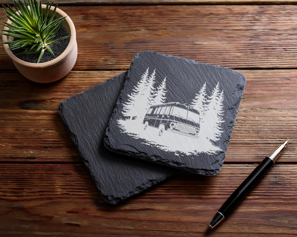 Slate coaster with engraved RV and pine trees, next to a pen and plant.