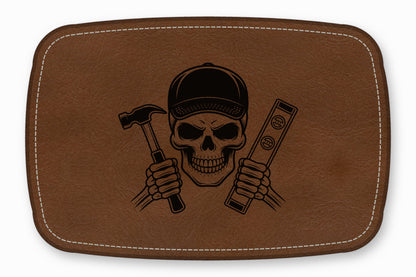 A brown leather patch features an engraved skull wearing a cap while holding a hammer and a level.