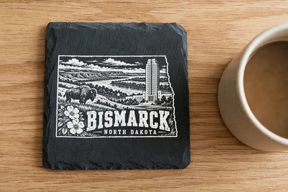 Square black slate coaster engraved with Bismarck, North Dakota landscape design alongside a coffee mug on wood surface.