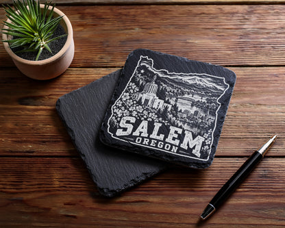 Two square black slate coasters on a wooden surface, one engraved with Salem Oregon and city landmarks.
