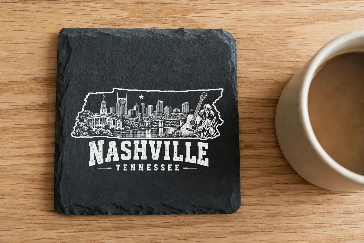 A square slate coaster engraved with a Nashville skyline graphic and text sits on a wooden table beside a coffee mug.