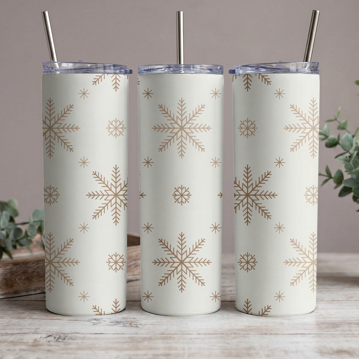 Three white tumblers with gold snowflake pattern and metal straws on a wooden surface.