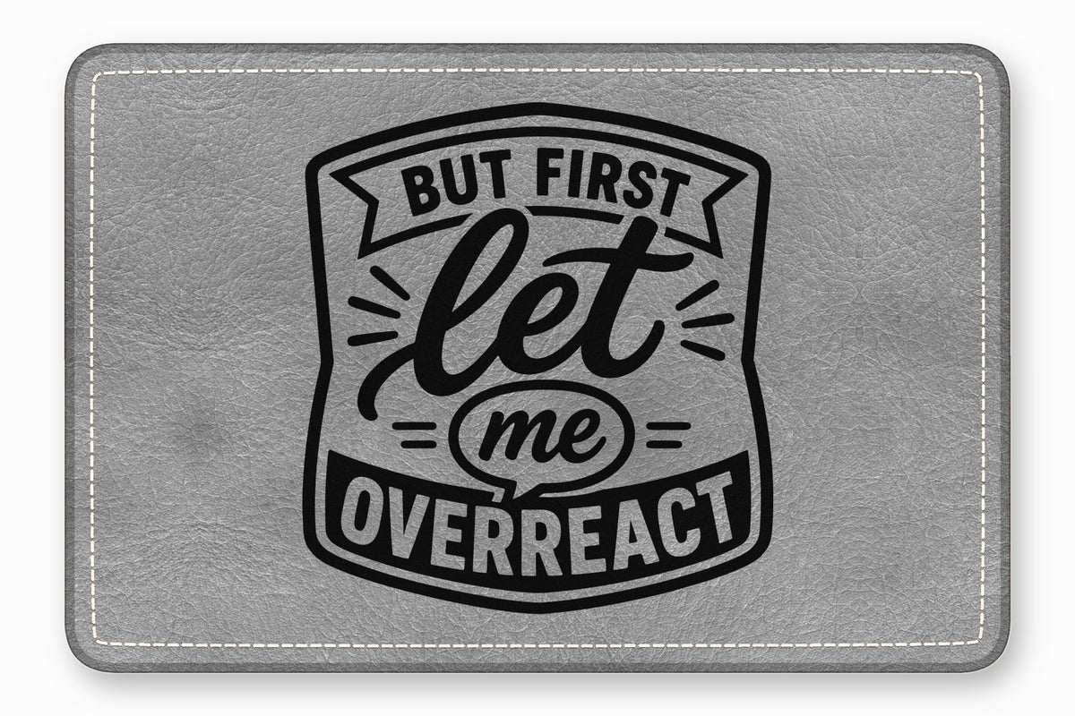 Gray leatherette patch with rounded corners, white stitching, and black engraved text reading But First Let Me Overreact.