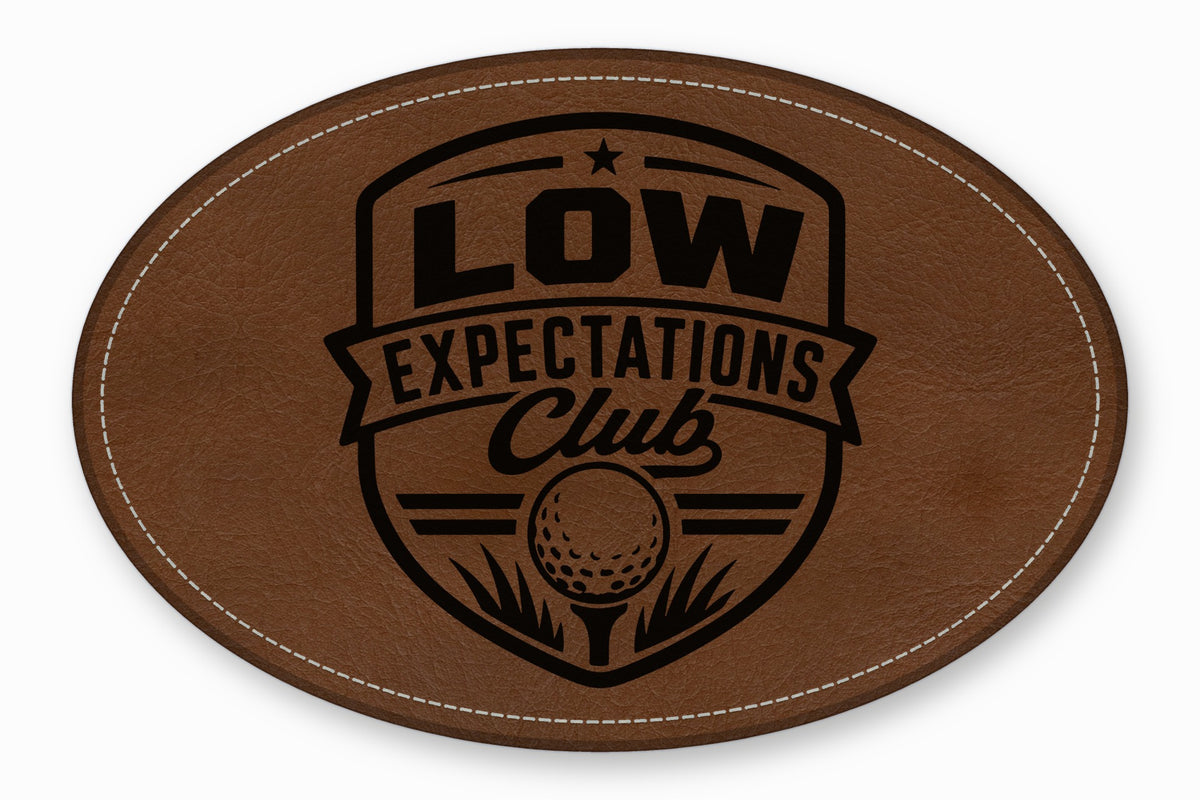 An oval brown leather patch with white stitched edges and a black golf-themed crest design reading Low Expectations Club.