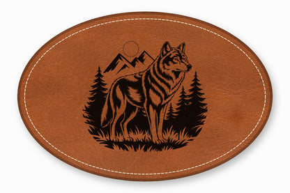 An oval brown leather patch featuring a laser-engraved design of a wolf standing before mountains and pine trees.