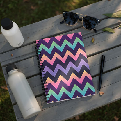 Spiral notebook with multicolor chevron pattern, water bottles, sunglasses, and pen on a wooden bench.