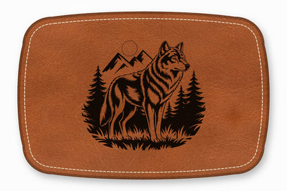 Brown leather rectangular patch with white stitched border and laser-engraved wolf, mountains, and pine trees.