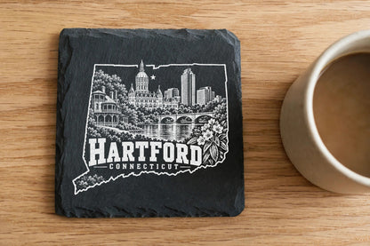 Square black slate coaster engraved with a Hartford Connecticut skyline design next to a coffee mug.