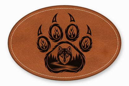An oval brown leather patch featuring a black engraved bear paw and a wolf head forest scene.