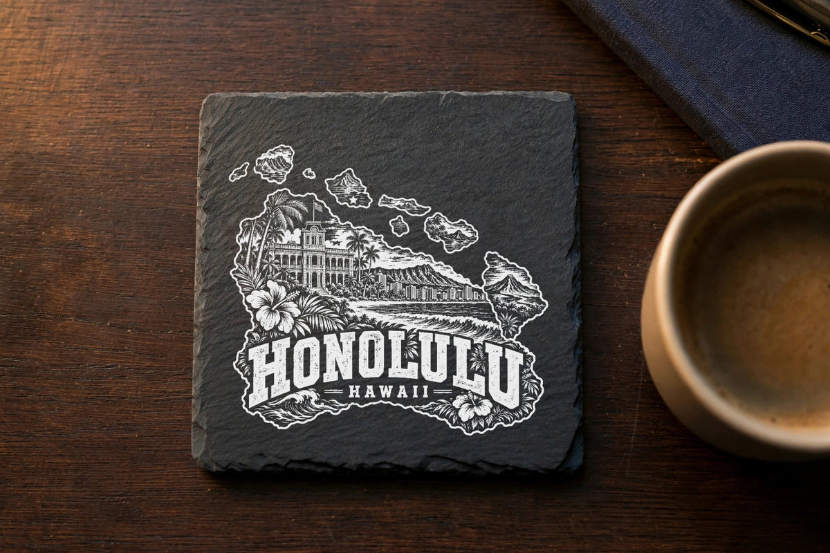 A square dark slate coaster engraved with a Honolulu Hawaii landscape design sits on a wooden table.