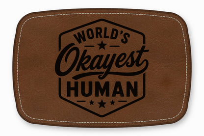 Brown rectangular leather patch with white stitched border, featuring the engraved text World's Okayest Human.