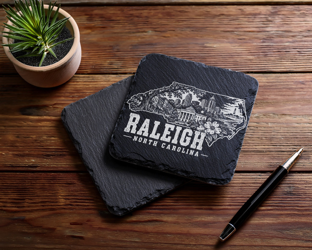 Two square black slate coasters engraved with a Raleigh, North Carolina cityscape design rest on a wooden table.