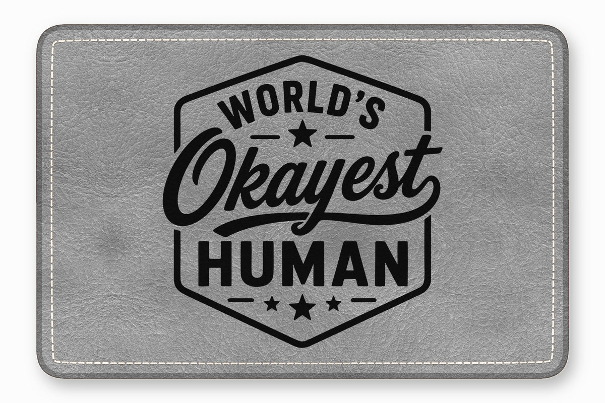 A rectangular gray faux leather patch with white stitching reads Worlds Okayest Human inside a hexagon.