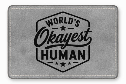 A rectangular gray faux leather patch with white stitching reads Worlds Okayest Human inside a hexagon.