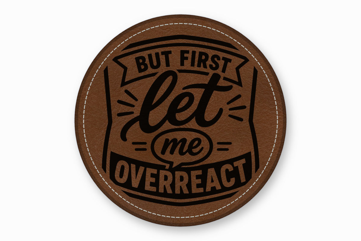 A round brown leather patch with white stitching and the engraved text But First Let Me Overreact.