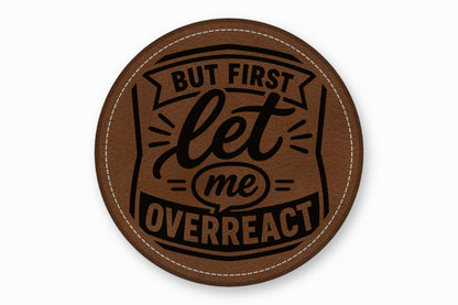 A round brown leather patch with white stitching and the engraved text But First Let Me Overreact.