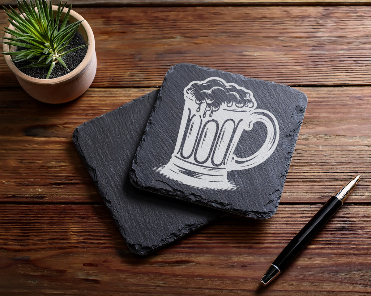 Slate coasters with etched beer mug design on wooden surface with pen and plant.