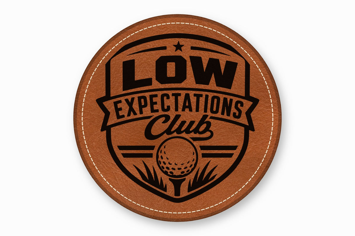 Round brown leather coaster featuring a black laser-engraved logo that reads Low Expectations Club with a golf ball.
