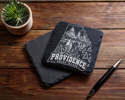 Two black square slate coasters rest on a wooden table alongside a potted succulent and pen.
